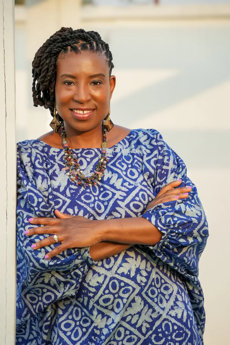 AmiCietta Duche Clarke, a smiling woman with braided hair wearing a blue patterned dress, leaning against a white wall in warm evening light.